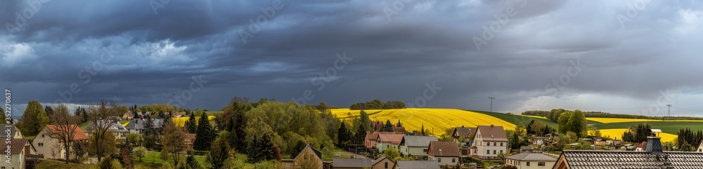 Panorama Dorf mit dahinterliegenden Feldern, betellt mit Raps unter dramatischem Himmel, Gewitterstimmung