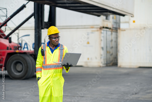 African technician dock worker in protective safety jumpsuit uniform and with hardhat and use laptop computer at cargo container shipping warehouse. transportation import,export logistic industrial
