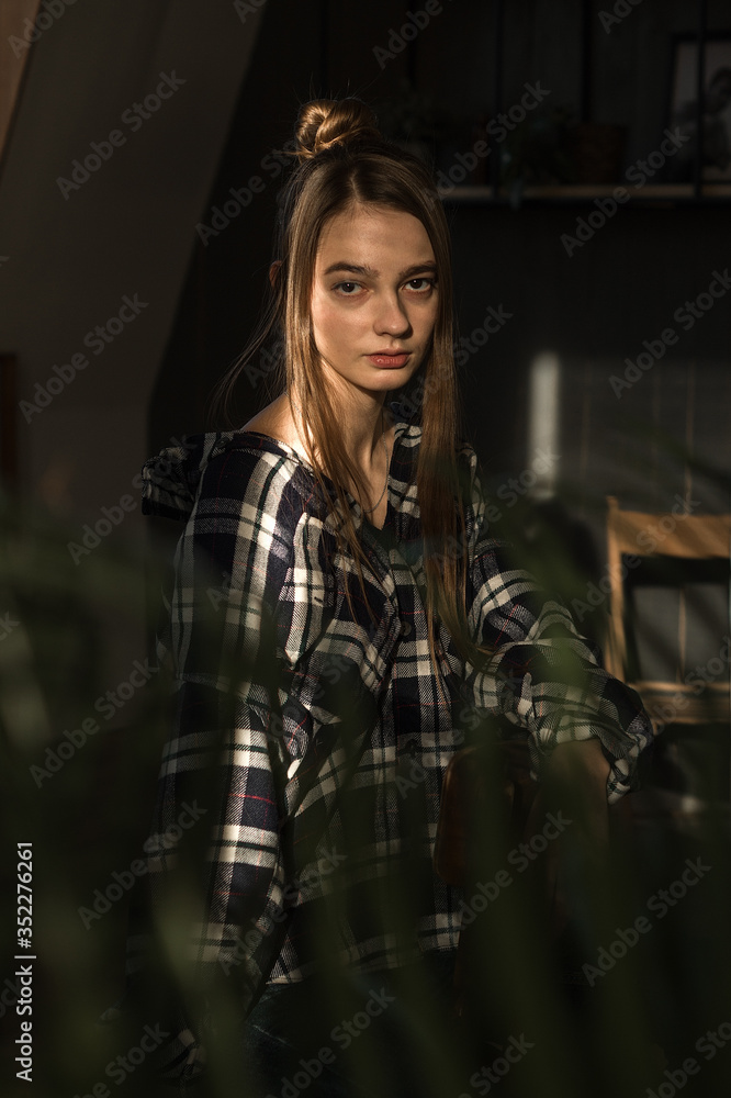 the girl sits on a chair in a plaid shirt, put her hands on the back of the chair, the ray of the sonde falls on her face the girl sits on a chair in a plaid shirt, put her hands on the back of the chair, the ray of the sonde falls on her face