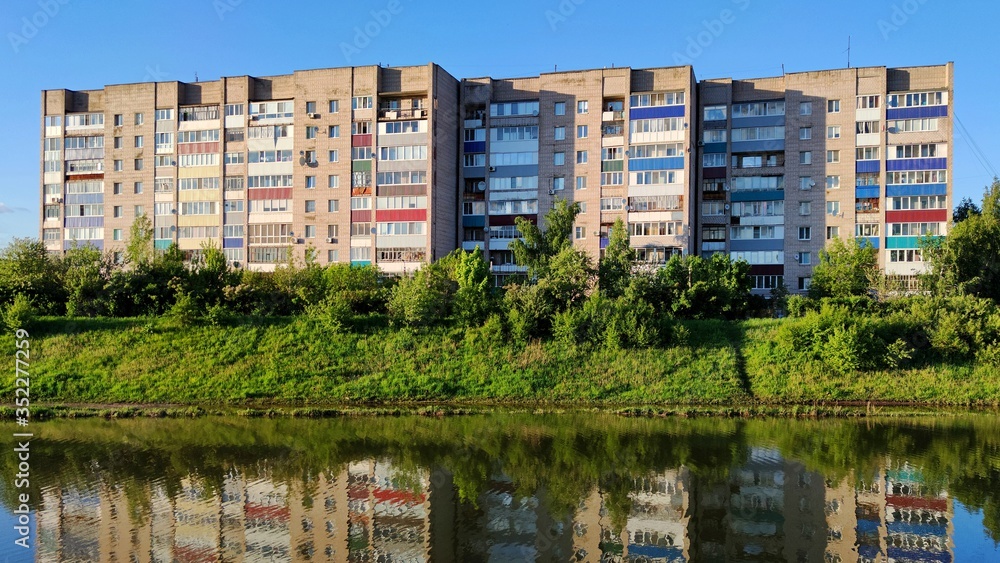 a multi-storey city house among green trees and grass is reflected in the water of the river against the blue sky in the rays of the evening sun