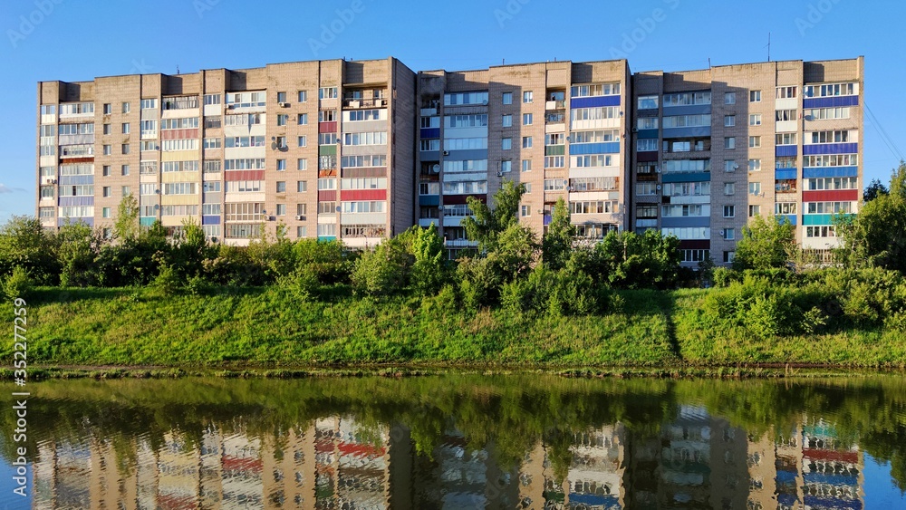 a multi-storey city house among green trees and grass is reflected in the water of the river against the blue sky in the rays of the evening sun a multi-storey city house among green trees and grass is reflected in the water of the river against the blue sky in the rays of the evening sun