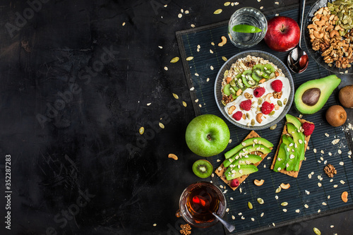 A healthy breakfast of cereals, fruits, berries, avocado, yogurt and nuts on a dark background. Healthy diet. A cup of tea. A glass with water. View from above. Space for text. Vegetarian menu.