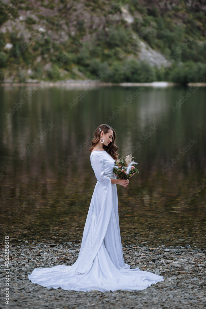 A bride in a long wedding dress standing on the shore of a lake A bride in a long wedding dress standing on the shore of a lake