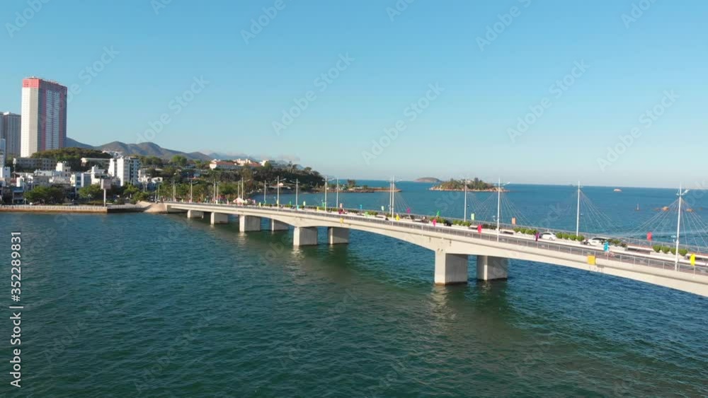 Shooting Aerial unmanned flight from top view of the city bridge with cars and motorbikes against the backdrop of the beautiful sea of the ocean and large town buildings. Bridge and city highway. Shooting Aerial unmanned flight from top view of the city bridge with cars and motorbikes against the backdrop of the beautiful sea of the ocean and large town buildings. Bridge and city highway.