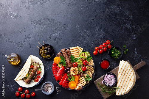 Grilled meat kebabs  vegetables on a black plate with tortillas  flat bread. Slate background. Copy space. Top view.