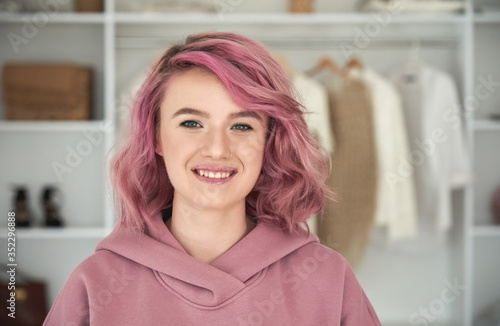 Happy hipster gen z teen girl fashion designer  stylist with pink hair and piercing wearing hoodie looking at camera in front of modern clothes wardrobe closet  face head shot close up portrait.