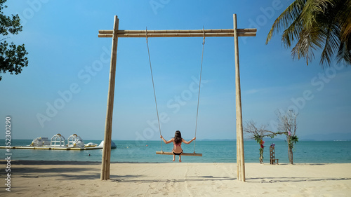 brunette lady with long hair swings on sand beach and enjoys landscape under green palms against blue sky backside view