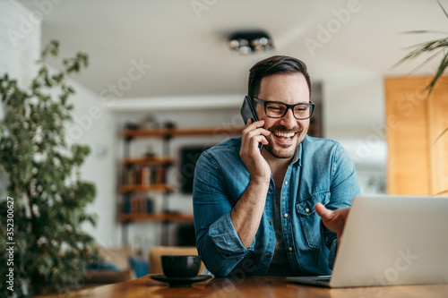Handsome smiling man talking on smart phone and looking at laptop at home, portrait.