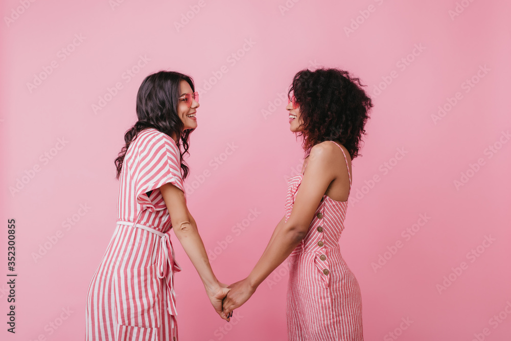 Cheerful brunette girls looking at each other while posing on pink background. Studio shot of lovely ladies in stylish summer attires Cheerful brunette girls looking at each other while posing on pink background. Studio shot of lovely ladies in stylish summer attires