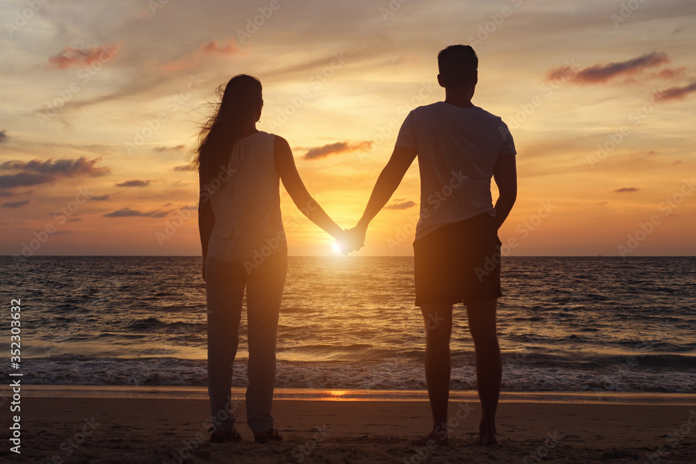 silhouette of young couple standing on beach to boundless ocean water joining hands at orange sunset backside view