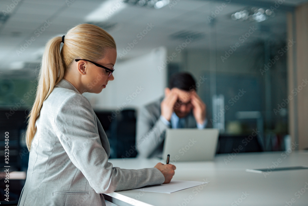 Portrait of a blonde businesswoman working, writing on paper. Coworker in the background is having problem. Portrait of a blonde businesswoman working, writing on paper. Coworker in the background is having problem.