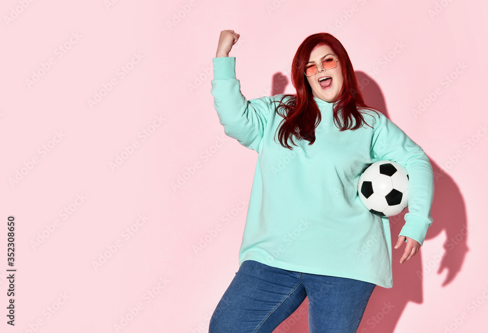 Young beautiful red haired overweight woman in jeans, blue hoodie and sunglasses standing with ball and expressing good luck Young beautiful red haired overweight woman in jeans, blue hoodie and sunglasses standing with ball and expressing good luck