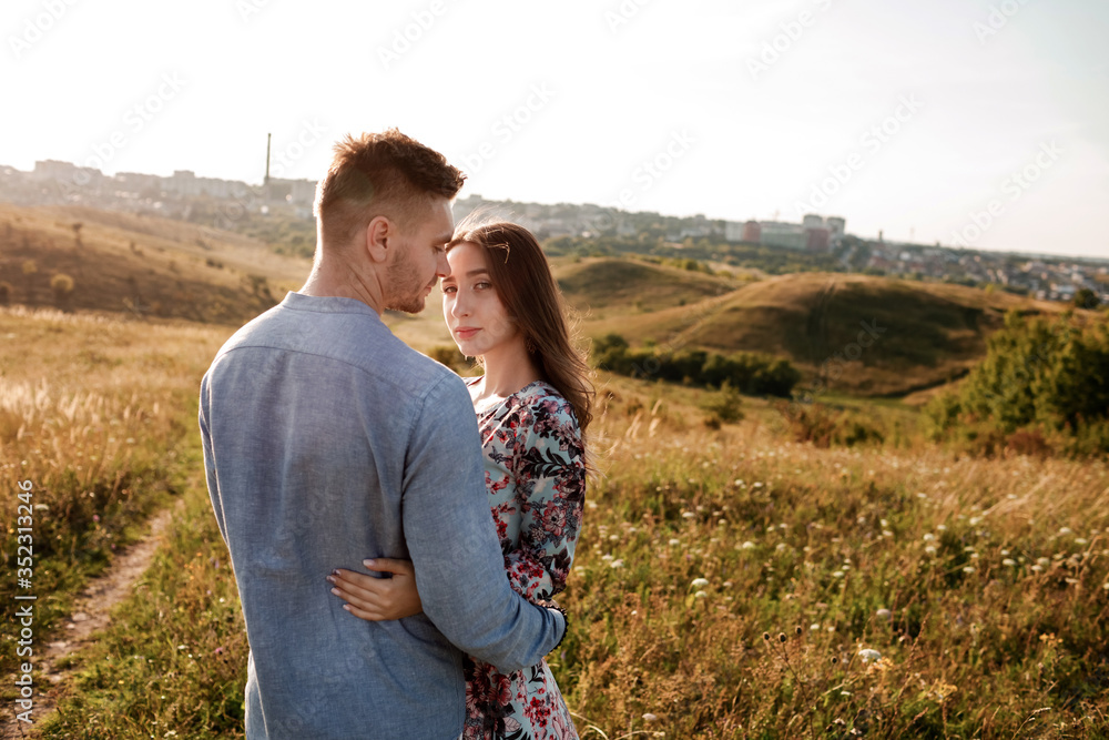 Young couple is hugging and walking in summer field with grass on the background town. Man and woman. Concept of lovely family. Young couple is hugging and walking in summer field with grass on the background town. Man and woman. Concept of lovely family.