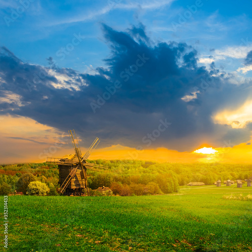 rural ethnic landscape, green fields with wooden windmill at the dramatic sunset