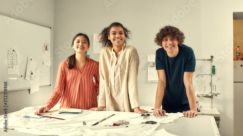 We are here for designing. Young cheerful designers smiling at camera while working together in a studio. Group of creative millennials measuring fabric, textile material before cutting it
