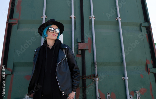 Girl with blue hair in a black hat and leather jacket poses near a green container.