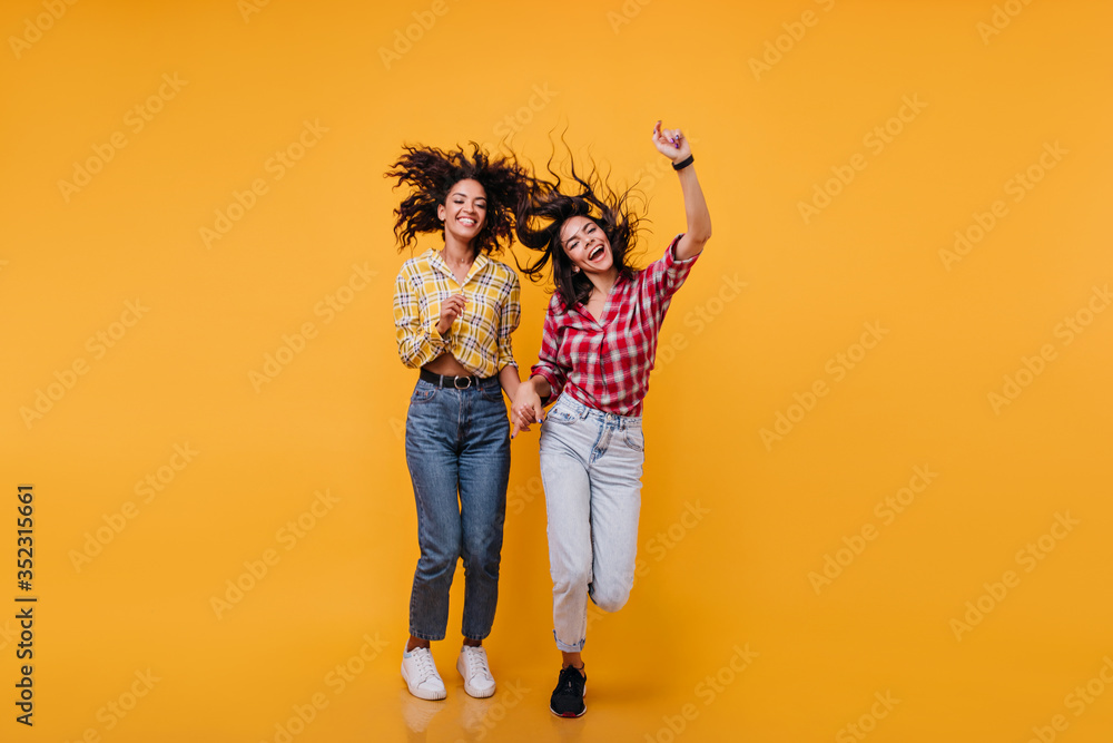 Positive girls jump on orange background. Full length shot of best friends in denim pants Positive girls jump on orange background. Full length shot of best friends in denim pants