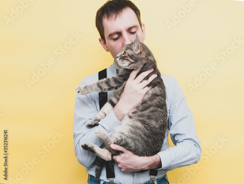 handsome man in blue shirt holding, hugging and kissing cute grey striped cat with outstretched paws on yellow background © Viktoriia