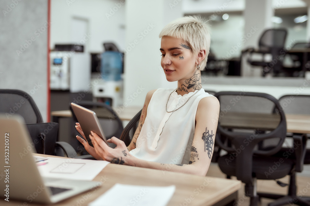Chatting with collegue. Young and attractive tattooed business woman using digital tablet and smiling while sitting at her workplace in the office. Business and career. Digital technologies at work Stock Photo | Chatting with collegue. Young and attractive tattooed business woman using digital tablet and smiling while sitting at her workplace in the office. Business and career. Digital technologies at work Stock Photo |