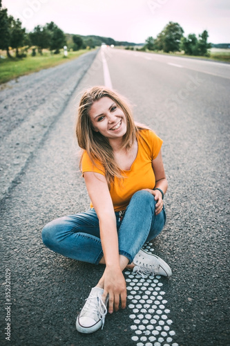  A young girl in a yellow T-shirt and blue jeans and white sneakers sits on the road and smiles. Evening, soft light. Light noise in the photo. Setting goals and moving towards them