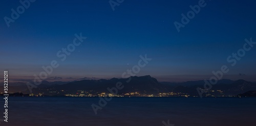 Night shot from a coast of Peschiera del Garda to the Garda lake and mountains with lights all over the other coast. Lights of Garda town in the night from other coast.