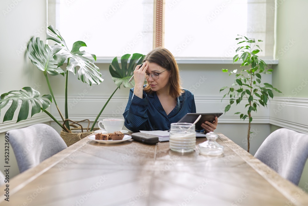 Serious mature woman working at home, using digital tablet, business notebook