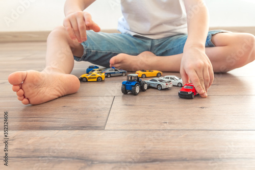 Little boy is playing with colored toy cars at home. Children feet barefoot on wooden warm floor. Joyful toddler sitting in room on laminate with  many favorite toys. Happy childhood
