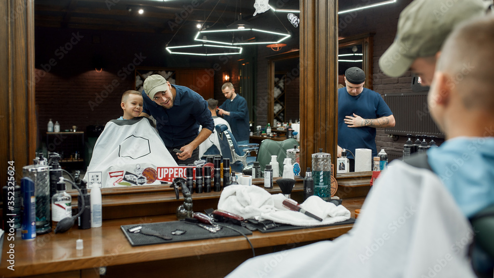 Friends. Young barber and cute little boy looking in the mirror at barbershop and smiling. Child visiting barber salon Friends. Young barber and cute little boy looking in the mirror at barbershop and smiling. Child visiting barber salon