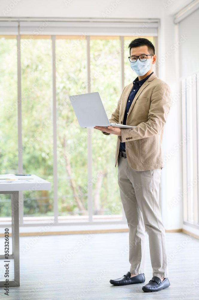 Fototapeta premium Happy Asian businessman entrepreneur wears face mask standing and holding laptop with confident in meeting room at office