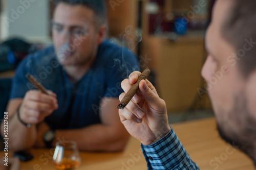 Side view of two friends sitting at the kitchen table and smoking Cuban cigarettes