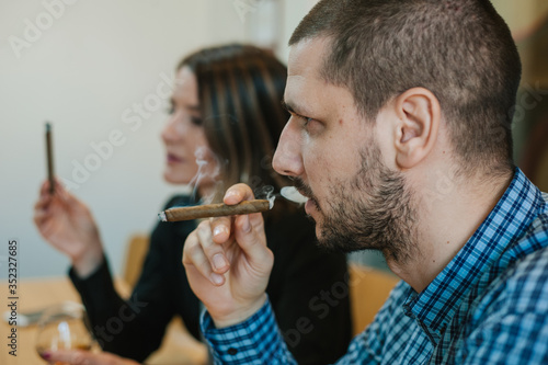 Side portrait of a white man with a beard smoking Cuban cigarettes. A woman in the background with a cigarette