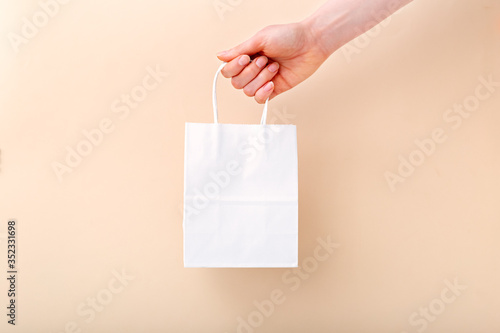 White paper shopping bag in female hand on beige light color background. Delivery, shopping, sale gift concept. Mockup bag packages. copy space for your text