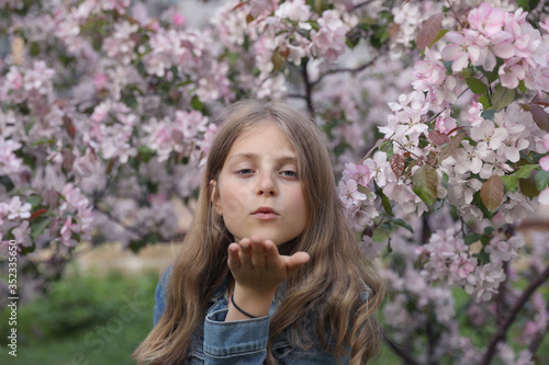 Portrait of a young girl in a flowering garden who sends an air kiss. The girl has long blond hair. The trees are beautiful pink flowers.