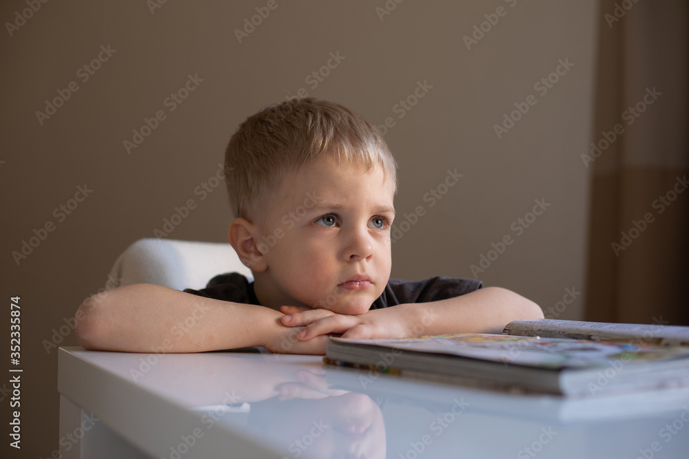 Portrait of a little boy with blond hair who sits at a table and puts his head on his hands Portrait of a little boy with blond hair who sits at a table and puts his head on his hands