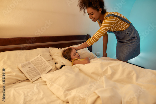 Every child is special. African american woman baby sitter spending time with little girl in the evening. Nanny tucking in kid, watching her falling asleep in bed