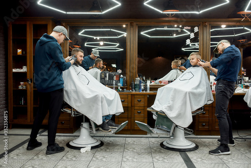 Family at barbershop. Young father and his little happy son sitting together in chairs in the modern barbershop while two barbers making haircuts for them photo