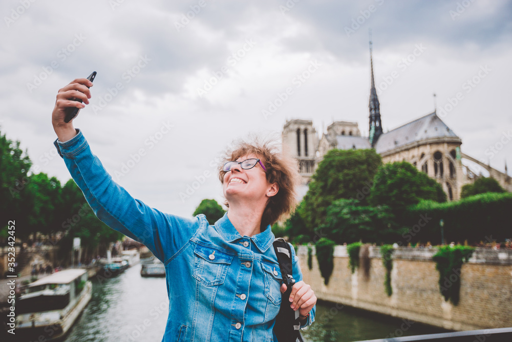 Fototapeta premium Caucasian female tourist photographing themselves by mobile telfon on background Notre Dame Cathedral from bridge over Seine bridge arsheveshe. Image Cathedral of Notre Dame de Paris