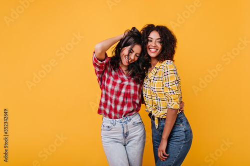 Emotional tanned women happily pose in plaid shirts against isolated background. Portrait of brunettes with curly hair © Look!
