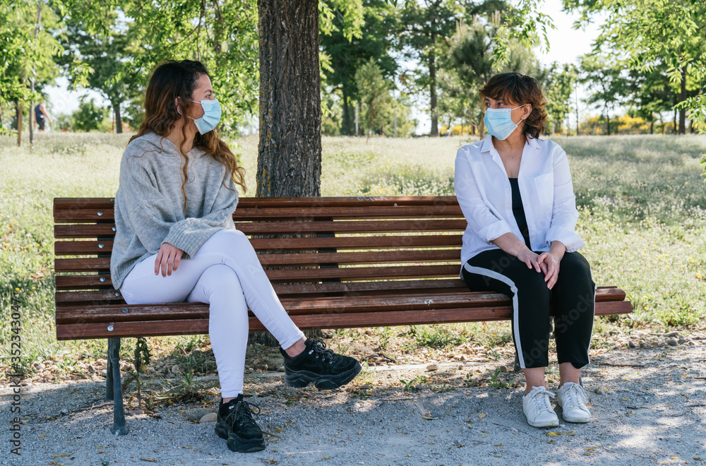 Social distance. Mother and daughter in social distance sitting on a park bench. Pandemic. Coronavirus. Maintaining safe distance sitting on a park bench. Safety distance. Social distance. Mother and daughter in social distance sitting on a park bench. Pandemic. Coronavirus. Maintaining safe distance sitting on a park bench. Safety distance.