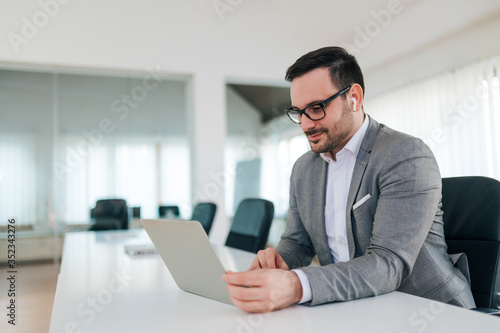 Portrait of a charming entrepreneur wearing suit and wireless earphones looking at laptop at board room.