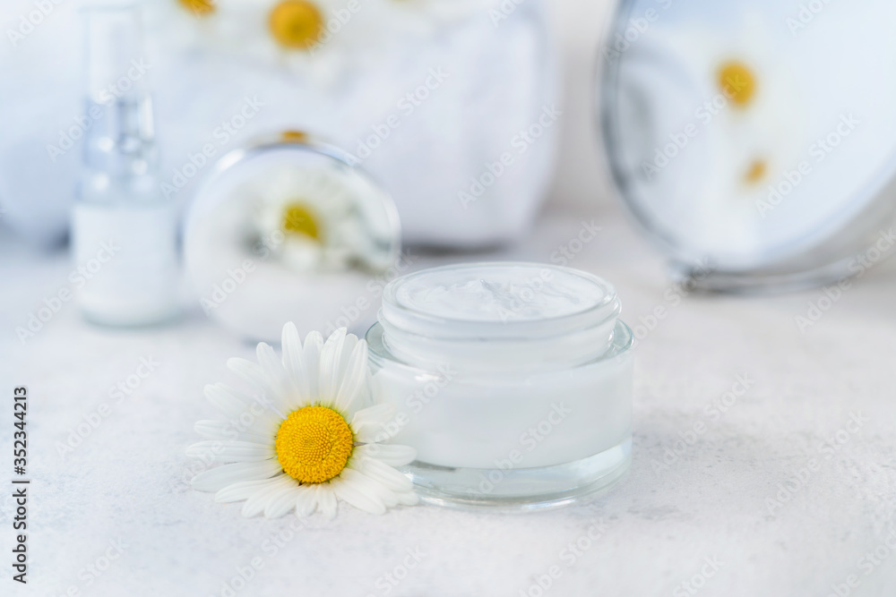 Herbal cosmetic cream in opened glass container and fresh chamomile flowers, towel, mirror on a white background. Natural organic moisturizer skincare product. Selective focus. Copy space. Herbal cosmetic cream in opened glass container and fresh chamomile flowers, towel, mirror on a white background. Natural organic moisturizer skincare product. Selective focus. Copy space.