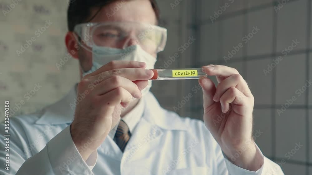A doctor in a protective mask holds a test tube with the COVID-19 virus in his hands and examines it. Close-up of a test tube with the virus. A doctor in a protective mask holds a test tube with the COVID-19 virus in his hands and examines it. Close-up of a test tube with the virus.
