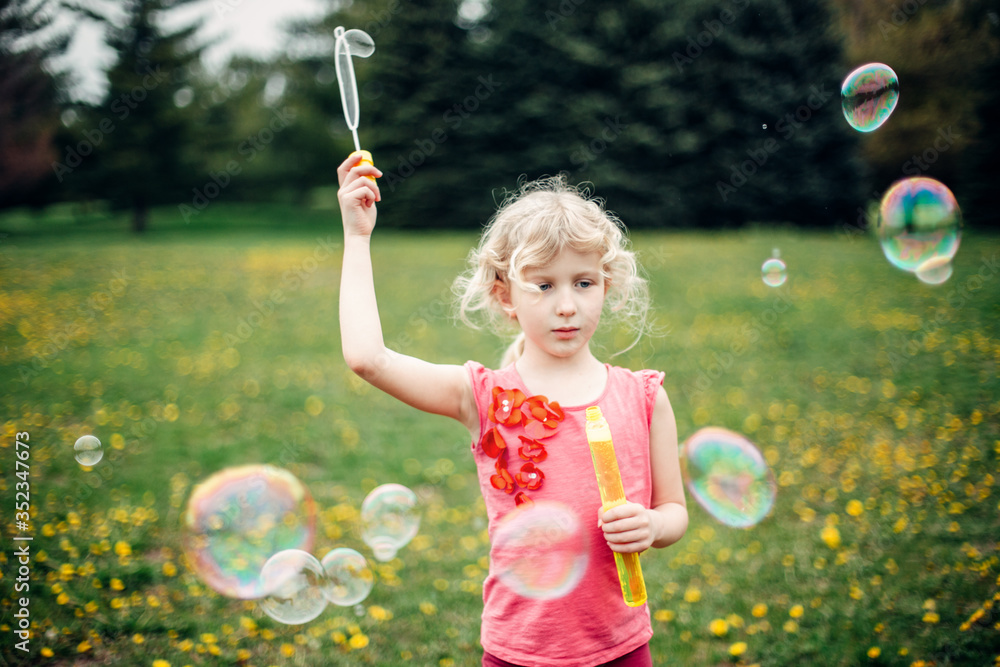 Preschool Caucasian blonde girl blowing soap bubbles in park on summer day. Child having fun outdoors. Authentic happy childhood magic moment. Lifestyle seasonal activity for children.