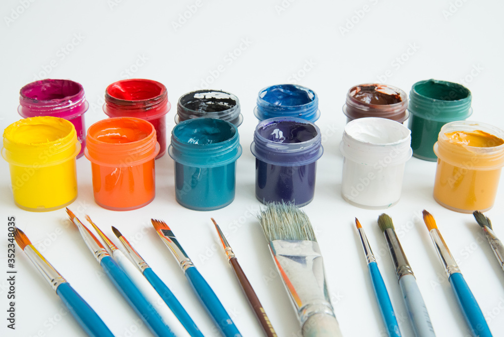 Jars of gouache paint in different colors. Next to it are brushes of various sizes. Artist's kit. Side view. White background. Hobbies and inspiration. Jars of gouache paint in different colors. Next to it are brushes of various sizes. Artist's kit. Side view. White background. Hobbies and inspiration.
