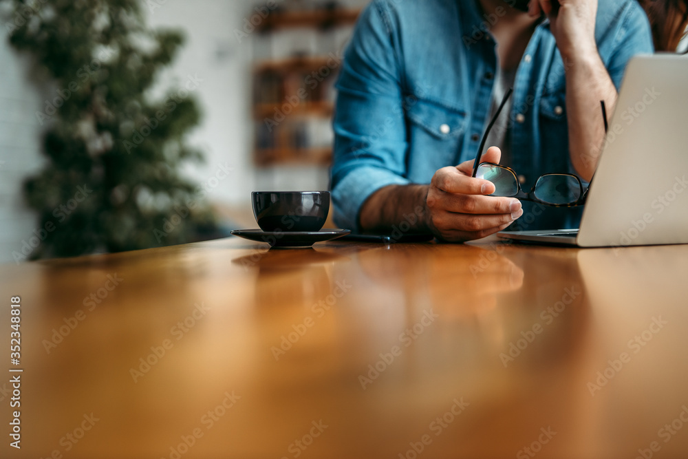 Close-up image of a man sitting at wooden table, copy space. Holding eyeglasses and looking at laptop. Close-up image of a man sitting at wooden table, copy space. Holding eyeglasses and looking at laptop.