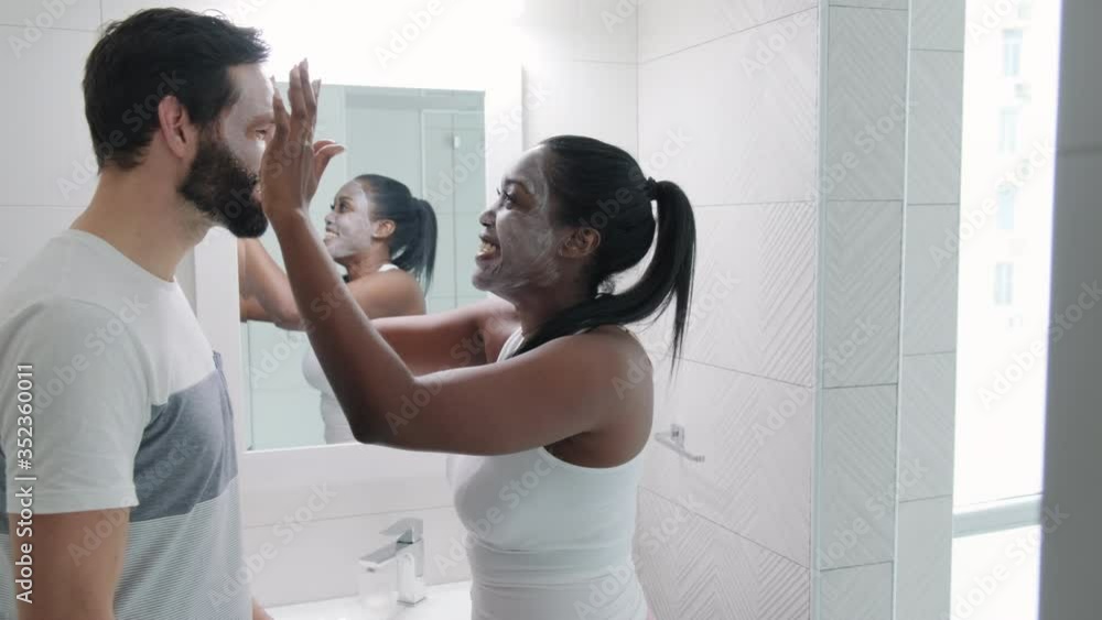 Multiethnic couple smiling and laughing in bathroom at home. Happy black woman applying beauty mask to white man. Married life and skin health for partners using cleansing lotion on face Multiethnic couple smiling and laughing in bathroom at home. Happy black woman applying beauty mask to white man. Married life and skin health for partners using cleansing lotion on face