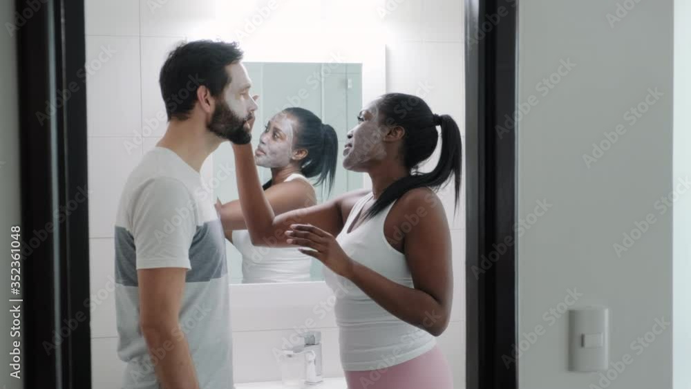 Interracial couple smiling and kissing in bathroom at home. Happy African American wife applying beauty mask to Caucasian husband. Married life and skin health for people using cleanser on face Interracial couple smiling and kissing in bathroom at home. Happy African American wife applying beauty mask to Caucasian husband. Married life and skin health for people using cleanser on face