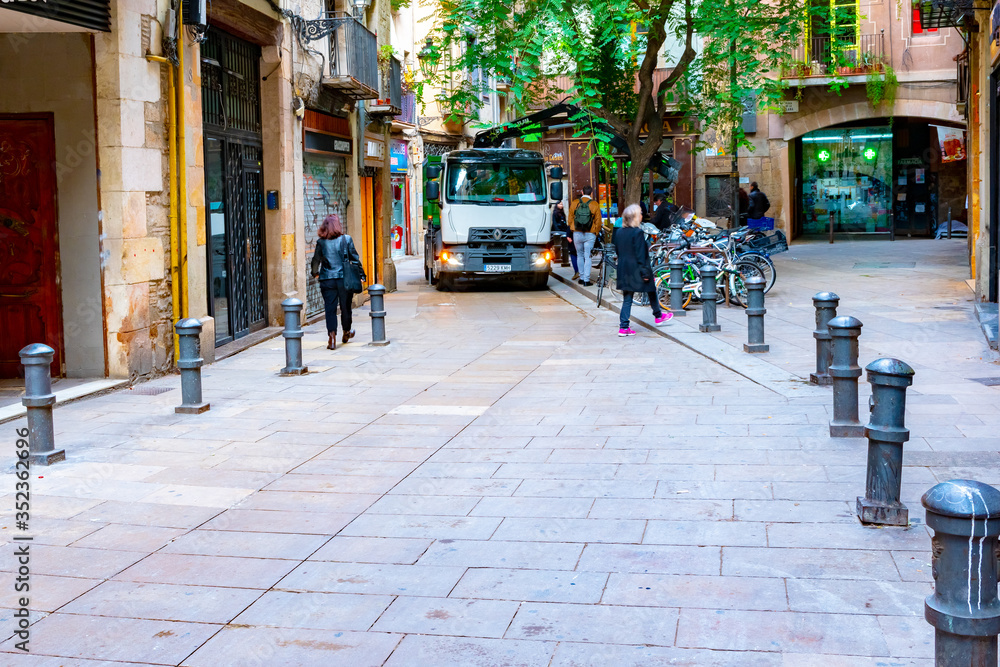 Barcelona, Spain. Truck drives down narrow street between stores in downtown Catalonia. People walk around bollard barriers and row of parked bicycles in commercial district. Barcelona, Spain. Truck drives down narrow street between stores in downtown Catalonia. People walk around bollard barriers and row of parked bicycles in commercial district.