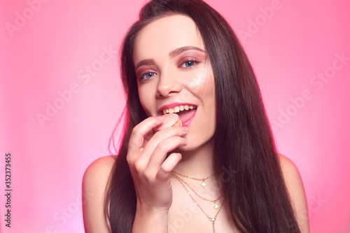 Girl eats macaroon, model on a pink background eats a sweet, sugar diet. Brunette with pink make-up, addicted to food.