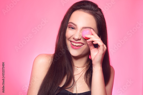 Girl eats macaroon, model on a pink background eats a sweet, sugar diet. Brunette with pink make-up, addicted to food.Girl eats macaroon, model on a pink background eats a sweet, sugar diet.